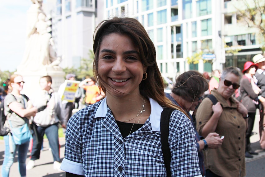 A teenage girl in a school uniform smiles in a crowded park.