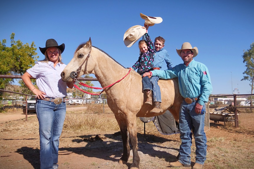 A father and mother wearing cowboy hats stand either side of a buckskin horse. Their children sit atop the horse.