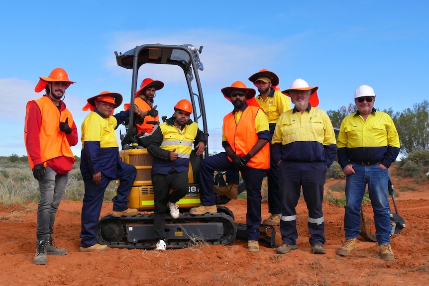A group of eight men in high-vis standing around an excavator.