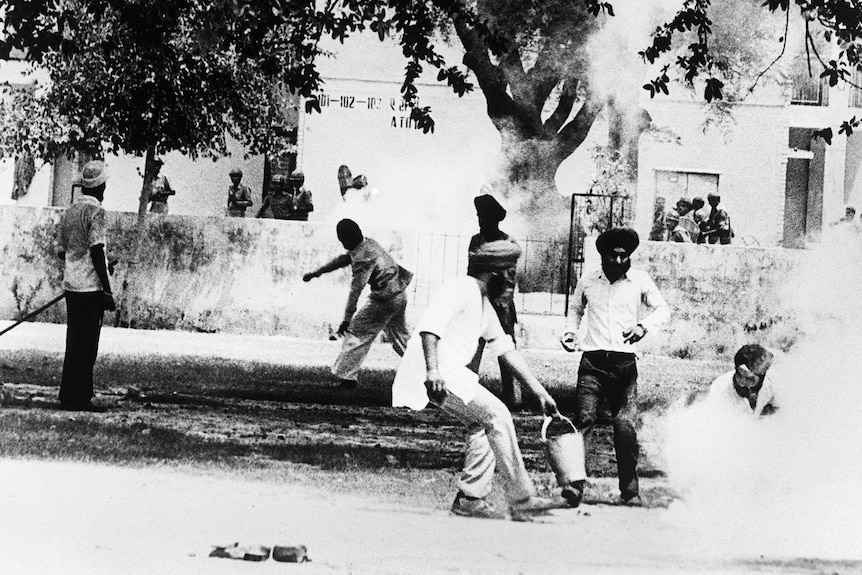 A black-and-white photo shows a man holding a tear gas canister, people run behind him with smoke in the air