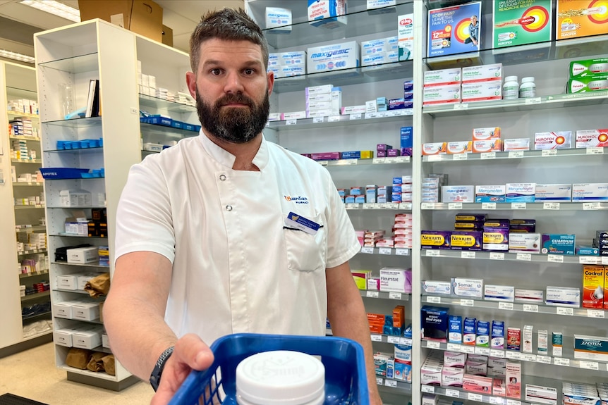 a pharmacist standing behind a counter holding a prescription basket towards the camera