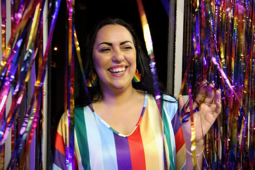 A lady is smiling, wearing a rainbow coloured dress and earrings, standing amongst glittery streamers. 