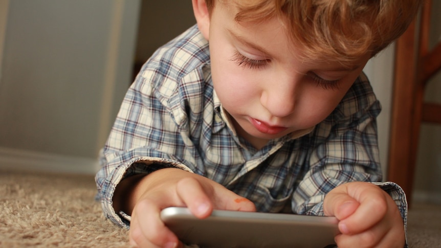 A young child lies on the carpet using a smartphone.