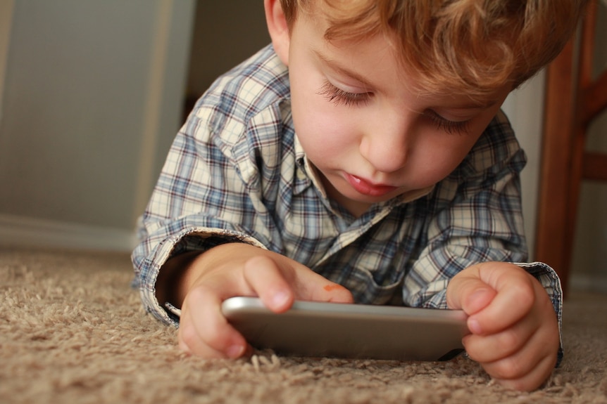 A young child lies on the carpet using a smartphone.