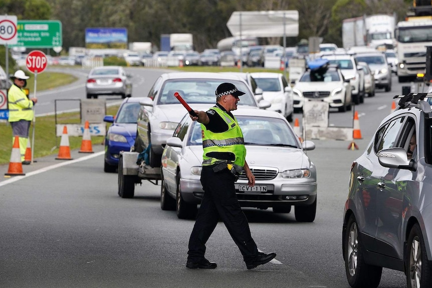 A police officer wearing a yellow hi-vis vest directs a long queue of traffic on a busy road.