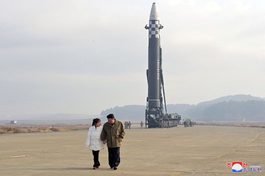 Kim Jong Un walks with his daughter on an open field with a missile behind him.
