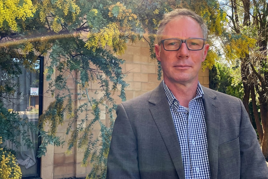 A man with short hair wearing glasses and a grey blazer looks seriously at the camera.