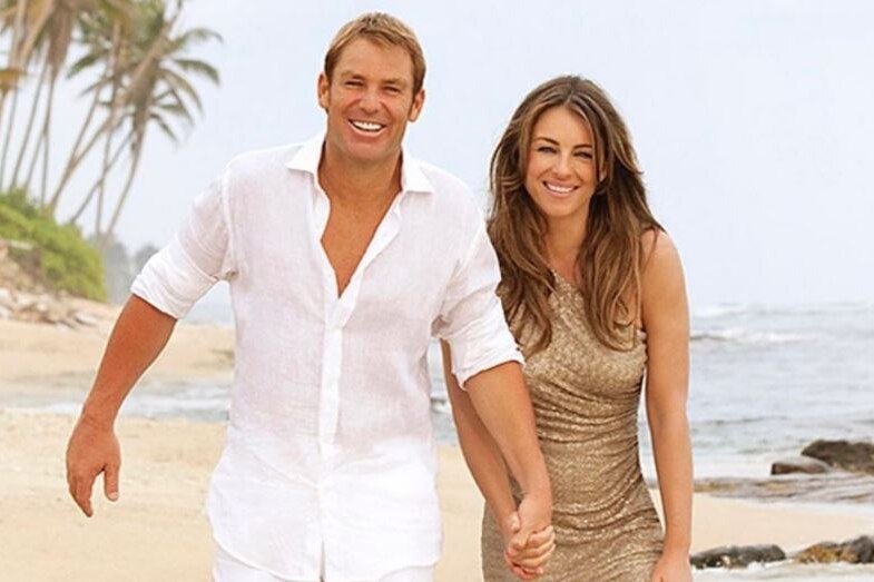 Shane Warne wears white and holds the hand of Elizabeth Hurley, in a gold dress, as they walk along the beach smiling.