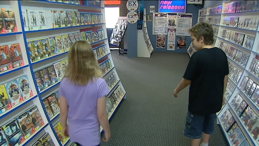 Two children walking through a Video Ezy video rental store aisle.