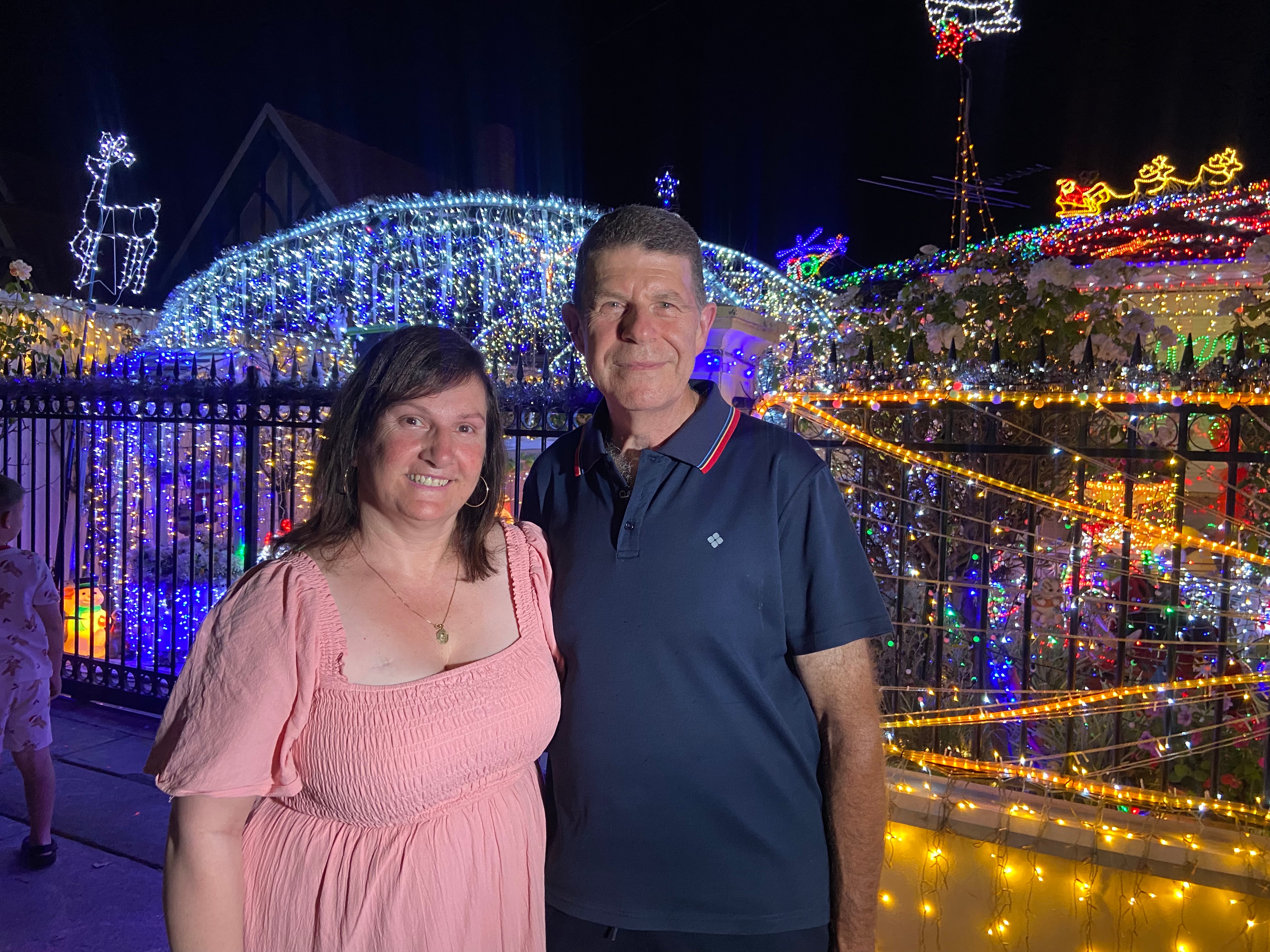 Uma mulher de vestido rosa e um homem de camisa azul posam para uma foto do lado de fora de uma casa decorada com luzes de Natal.