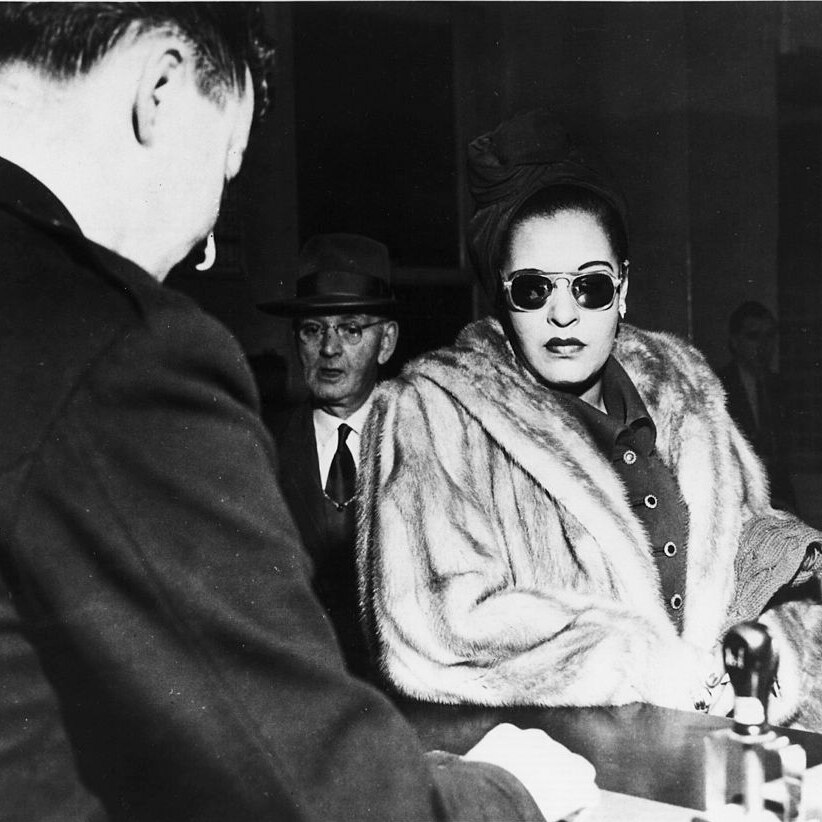 Wearing dark sunglasses and fur coat,  Billie Holiday stands before a desk, surrounded by suited white men.