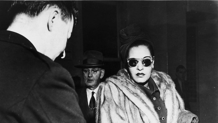 Wearing dark sunglasses and fur coat,  Billie Holiday stands before a desk, surrounded by suited white men.