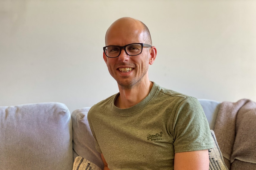 A man in glasses and a t-shirt smiling on his couch.