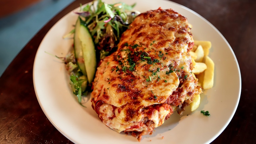 A chicken parmigiana sits atop chips and a side salad on a white plate, which is on a wooden bench