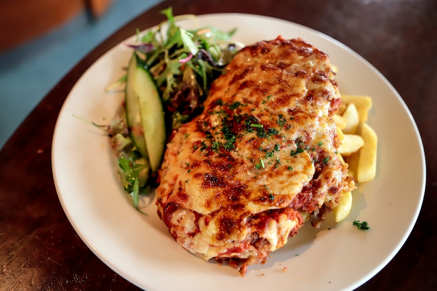 A chicken parmigiana sits atop chips and a side salad on a white plate, which is on a wooden bench