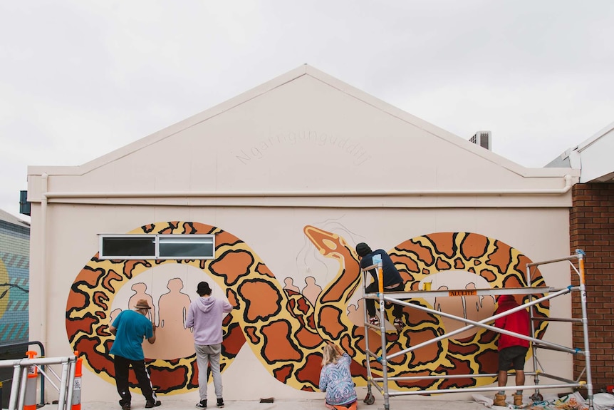 A large snake mural being painted on a wall by 4 people.