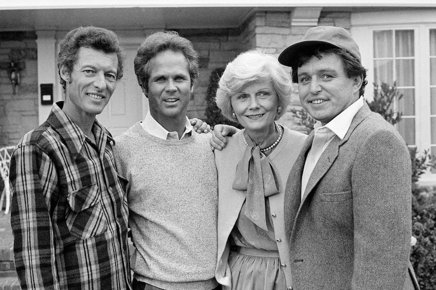A black and white photo of four actors dressed in 1950s clothing smiling in front of a brick suburban house.