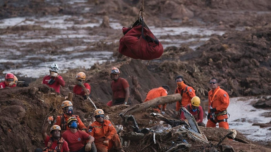 A group of Brazilian rescuers watch a body being airlifted in a red bag in a field with mounds of brown mud and dirt.