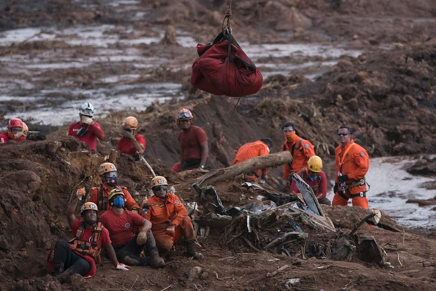 A group of Brazilian rescuers watch a body being airlifted in a red bag in a field with mounds of brown mud and dirt.