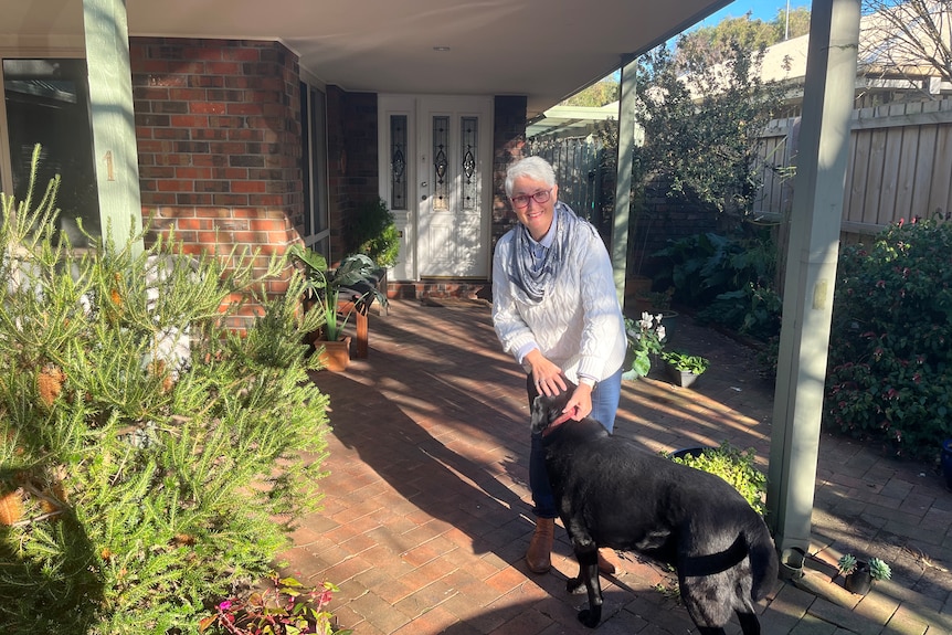 A smiling, older woman outside her house patting a black labrador.