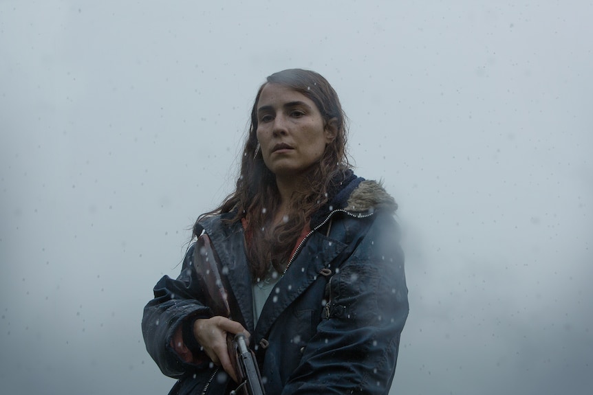 Woman with wet dark hair, wearing a navy hooded jacket and holding a shotgun is shrouded in fog and rain.