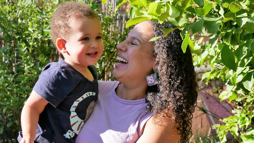 woman holding her two year old child, smiling, in front of green bush