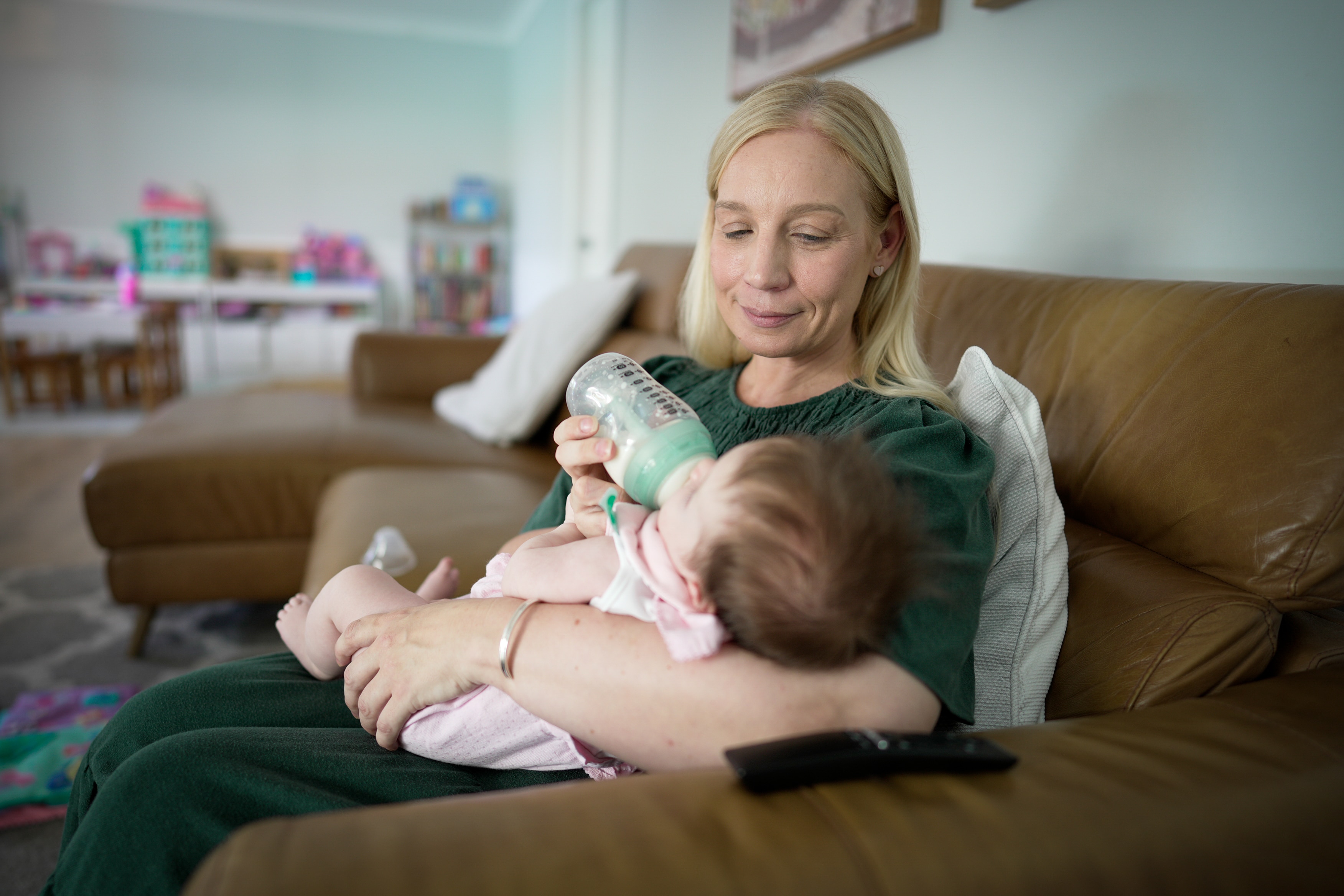 A woman sits on a couch feeding a bottle to a baby.