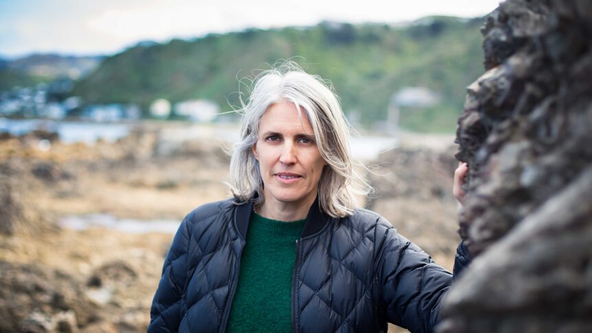 Ingrid Horrocks stands near rockpools with water and hills in the distance. She has white hair and looks intently at the camera.