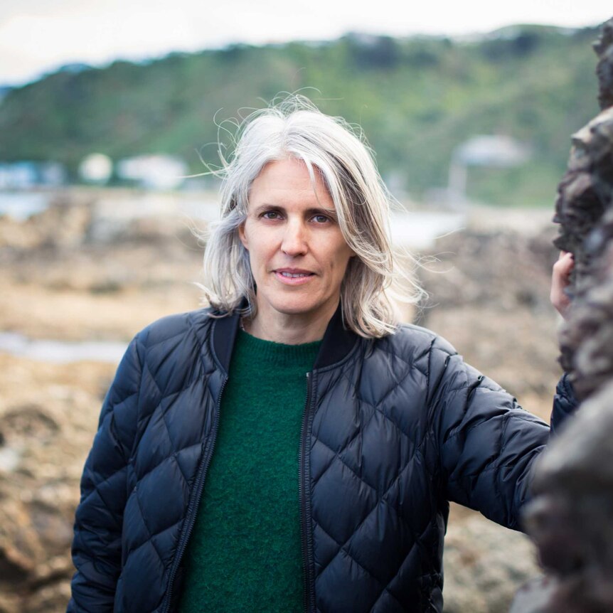 Ingrid Horrocks stands near rockpools with water and hills in the distance. She has white hair and looks intently at the camera.