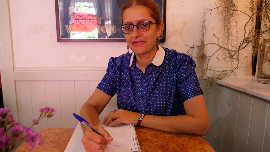 A woman in a blue polka dot shirt with glasses holds a pen above an A4 notepad. 