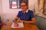 A woman in a blue polka dot shirt with glasses holds a pen above an A4 notepad. 