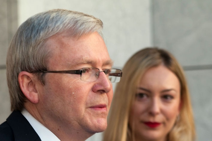 A tear slides down Kevin Rudd's face as he addresses a press conference. A blonde woman watches him.