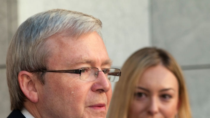 A tear slides down Kevin Rudd's face as he addresses a press conference. A blonde woman watches him.