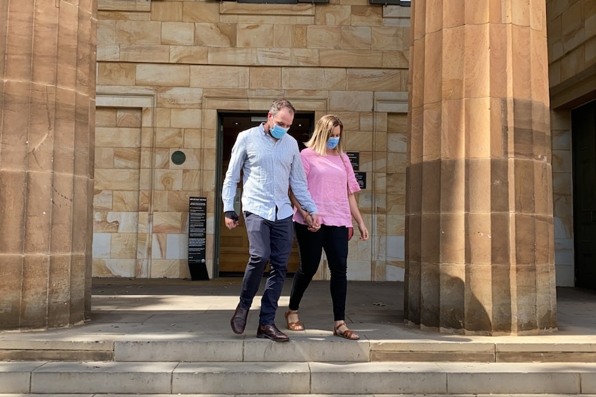 A man in navy pants and a blue shirt holds the hand of a woman in a pink shirt and jeans as he walks out of court