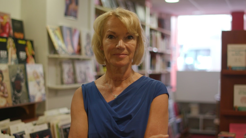 Brigitte Lahaie looks at the camera with a small smile, standing in a room with shelves in the background