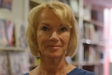 Brigitte Lahaie looks at the camera with a small smile, standing in a room with shelves in the background