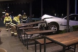 Firefighters in yellow uniforms and hard hats near a damaged white sedan stopped into outdoor dining tables