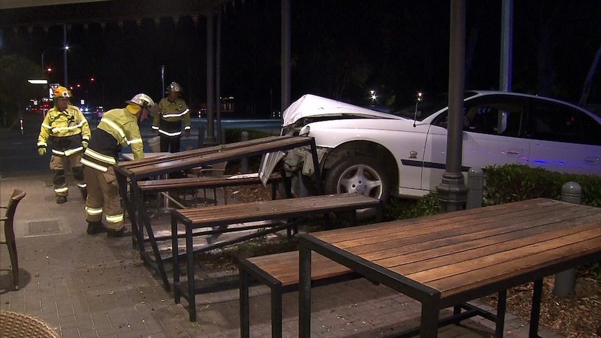 Firefighters in yellow uniforms and hard hats near a damaged white sedan stopped into outdoor dining tables