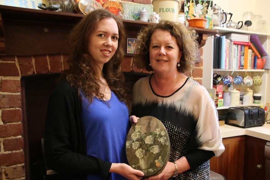 Matilda and her mother Kate holding the tin that belonged to Matilda's grandmother, in front of brick fire place