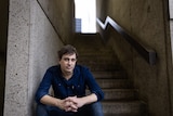 A middle-aged man with dark hair and wearing a navy shirt sits in a stairwell with his hands clenched together