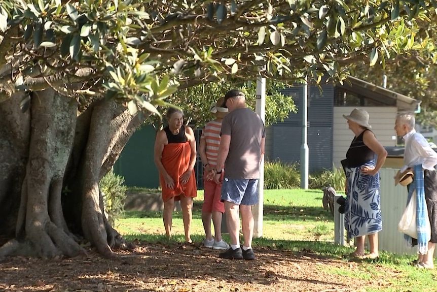 A group of people in bathers and towels talk near a tree.