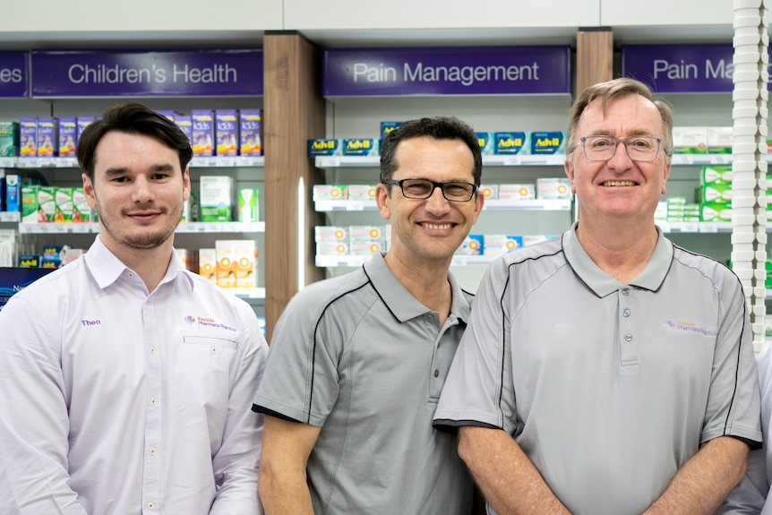 Three pharmacist in purple and grey uniform standing in front of medicines. 
