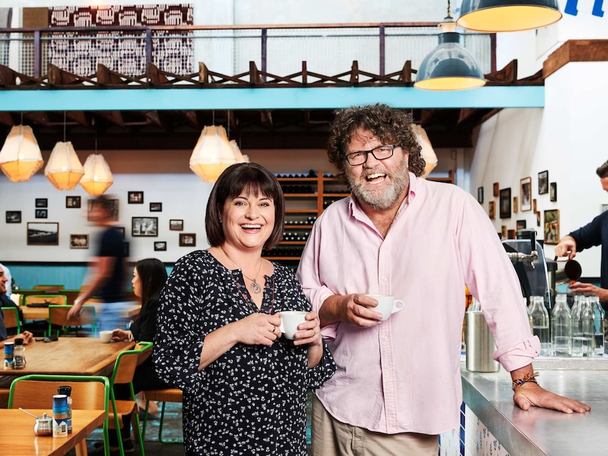 A woman with dark hair and a man with curly hair and a beard, standing in a cafe that has a loft area behind.