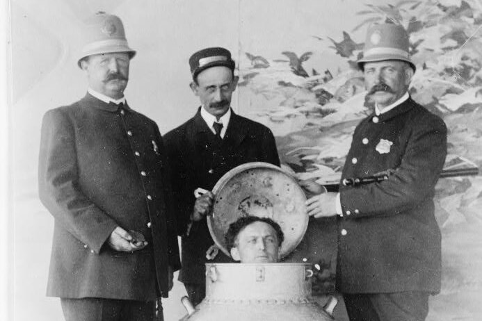 A man's head peeks out of a milk can as three police officers stand around him 