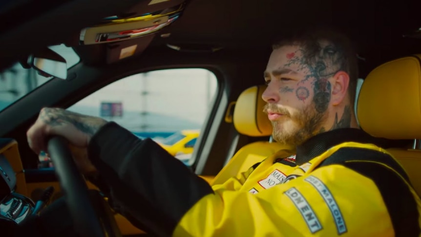 Post Malone sitting in the drivers seat of a care, wearing yellow and black jacket