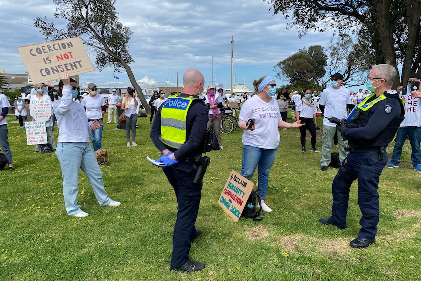 Two police officers stand with protesters who hold signs in a park field on a sunny day.