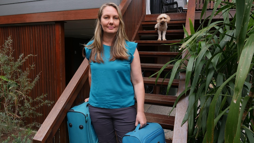 A woman in a blue shirt stands on a staircase holding two suitcases.