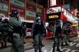 Riot police wearing face mask stand outside a shopping mall at night as a red bus drives by.