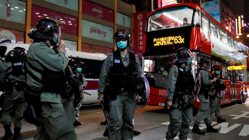 Riot police wearing face mask stand outside a shopping mall at night as a red bus drives by.