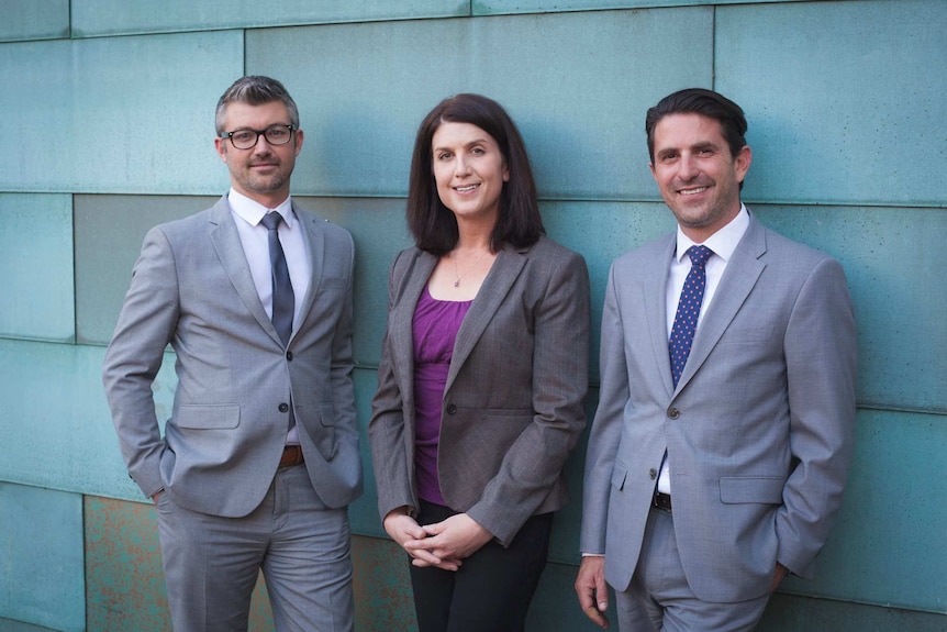 Three lawyers in suits, Autumn Scardina in the centre, stand in front of a wall with large blue tiles.